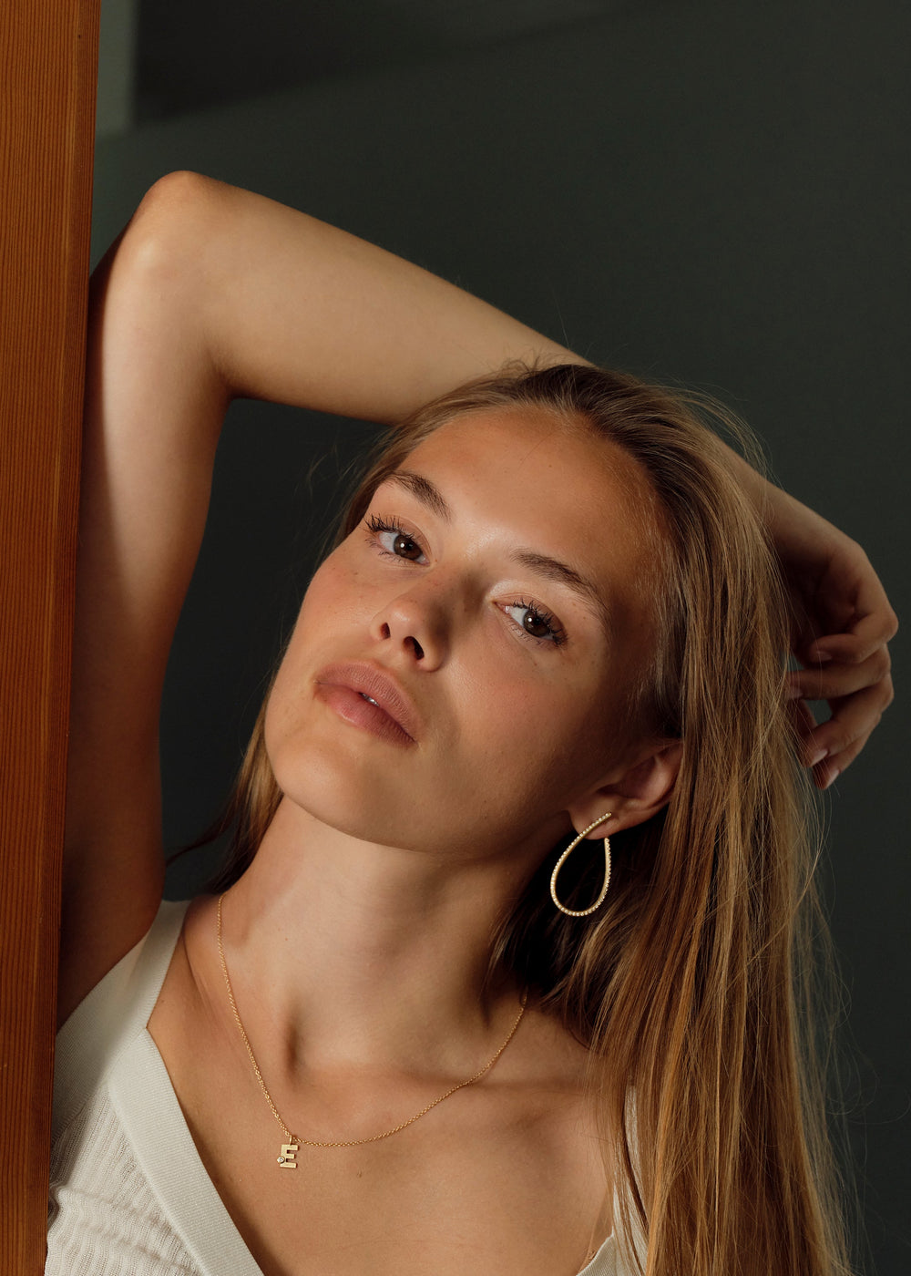 Woman with long hair leaning against a wooden wall indoors. Wearing big diamond earrings.